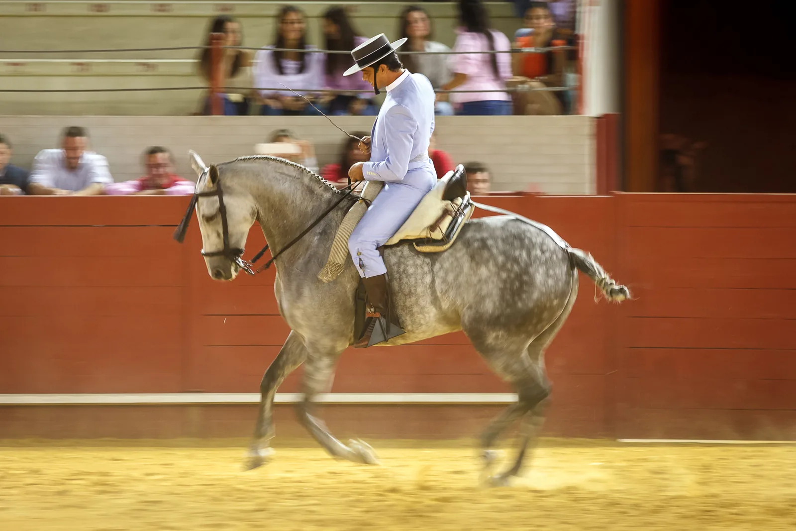 A rider in a short pale jacket and flat-brimmed hat on a dappled grey horse, cantering past a red arena wall with spectators watching from the stands behind